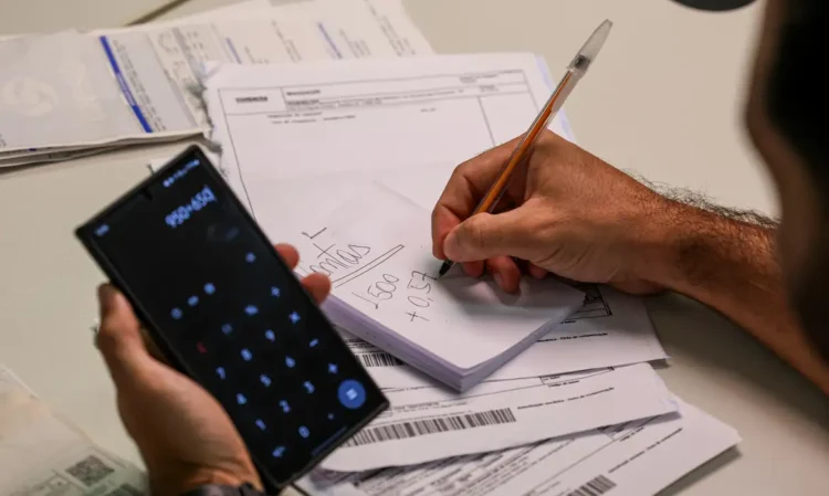 Pessoa usando calculadora no celular e escrevendo cálculos financeiros em bloco de notas sobre faturas