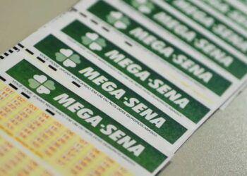 Close-up inclinado de vários volantes de aposta da Mega-Sena empilhados, mostrando o logotipo verde e os campos de números para marcação.