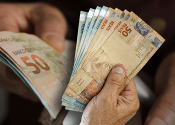 Mãos de pessoa contando várias cédulas de cinquenta reais (R$ 50,00), ilustrando o tema de finanças, dinheiro e o possível pagamento do 13º salário.