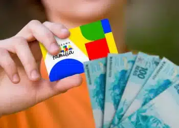 Cartão do Programa Bolsa Família sendo segurado com dinheiro em notas de 100 reais.