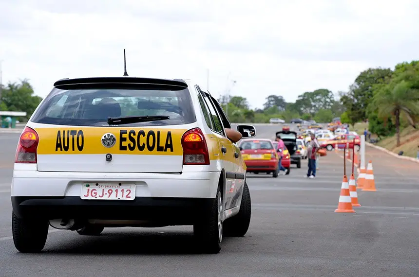 Carro de autoescola em treinamento prático. Carro de autoescola realizando teste em pista com cones e outros veículos à frente.
