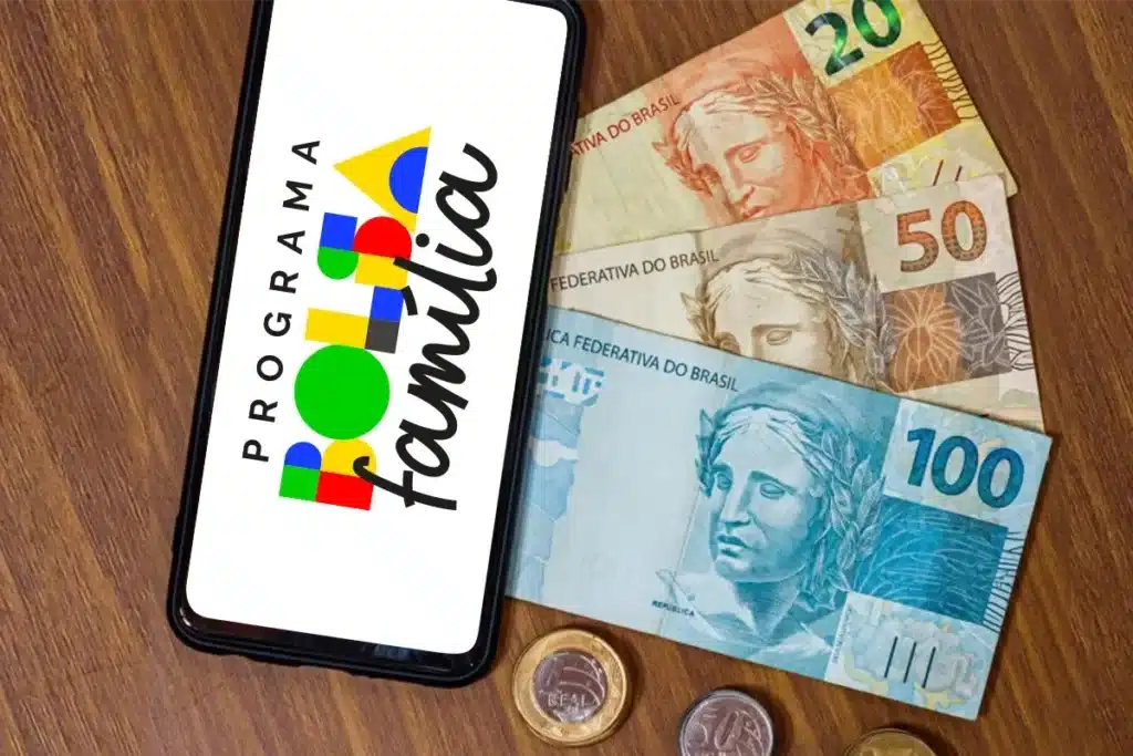 Benefício do Bolsa Família: cédulas e celular com aplicativo. Cartão do Bolsa Família exibido em um celular, com cédulas e moedas brasileiras ao fundo, ilustrando os benefícios.