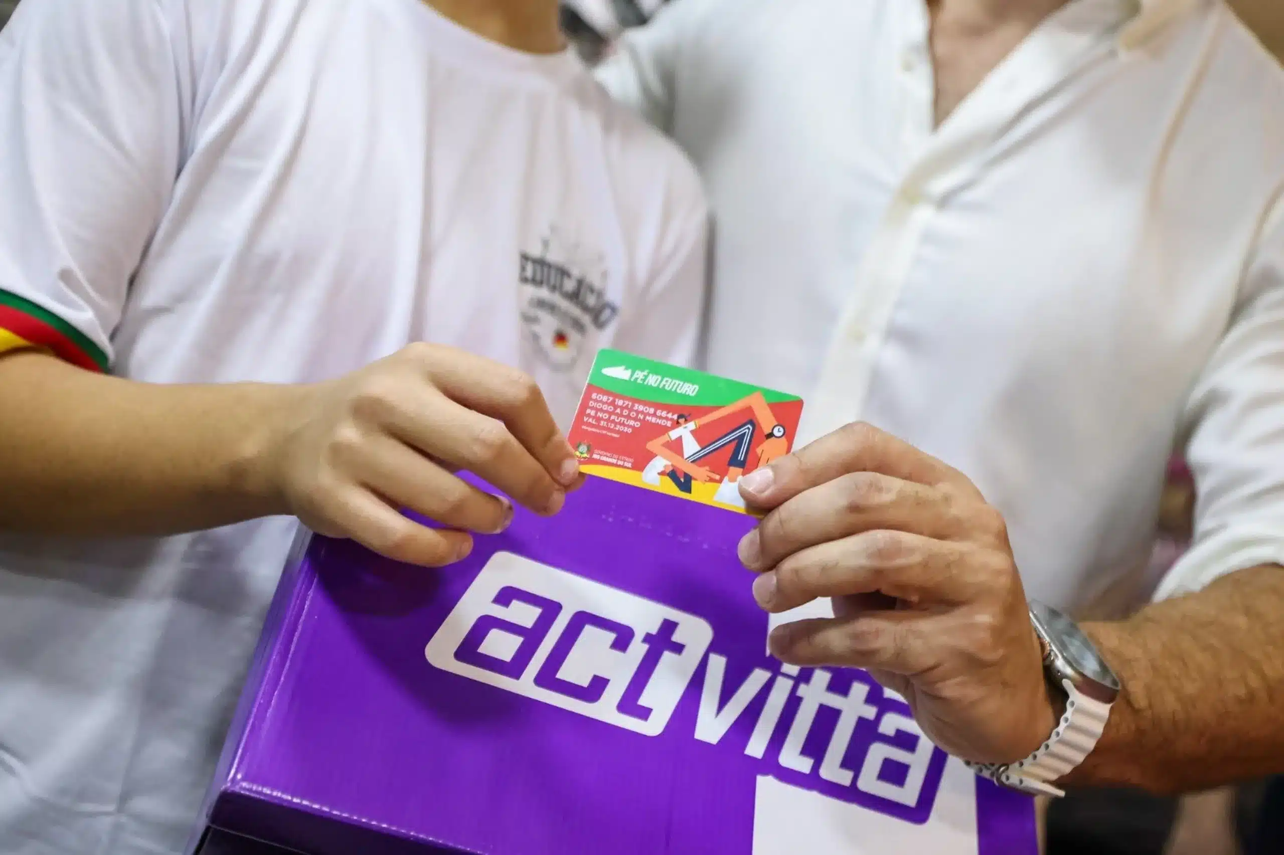 Programa Pé no Futuro. Programa Pé no Futuro, duas pessoas mostrando o cartão do programa sendo entregue a um estudante, simbolizando o auxílio financeiro para a compra de tênis e meias.