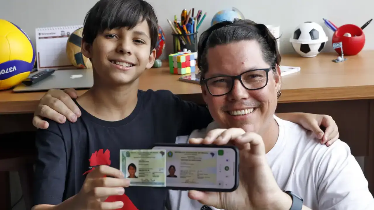 Pai e filho exibem documento digital em ambiente doméstico Homem de óculos e criança sorridente mostram celular com documento digital em frente à mesa com materiais escolares