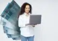 Mulher sorridente consulta notebook à frente de notas de 100 reais