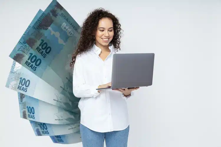 Mulher sorridente consulta notebook à frente de notas de 100 reais