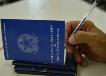 Mão segurando caneta preenche carteira de trabalho azul sobre mesa clara