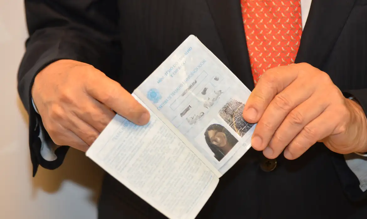 Carteira de Trabalho apresentada em ambiente corporativo Mãos masculinas segurando carteira de trabalho aberta com foto e identificação visíveis