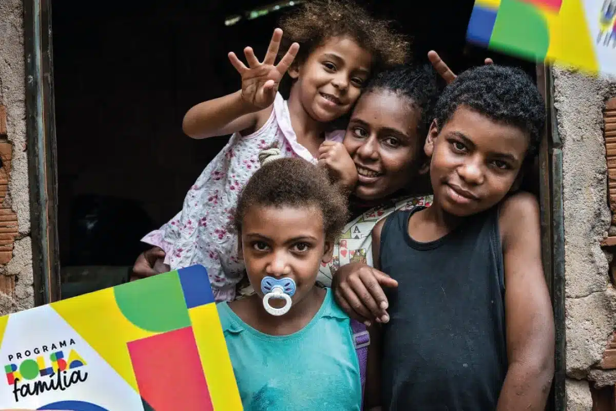 Crianças sorrindo em janela de casa beneficiadas pelo Bolsa Família 2026 Quatro crianças juntas na janela sorrindo, com cartão do Bolsa Família em destaque