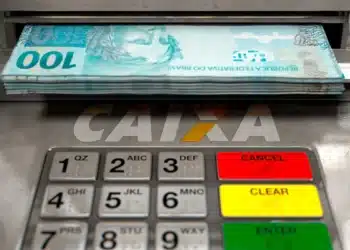 Entenda como sacar até R$ 6.220 da CAIXA pelo celular e sem sair de casa 8 Pilha de notas de 100 reais saindo de caixa eletrônico CAIXA com teclado em destaque.