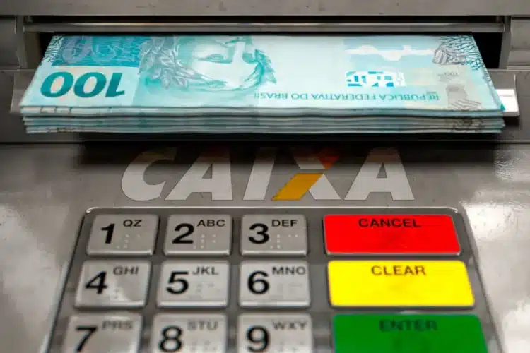 Pilha de notas de 100 reais saindo de caixa eletrônico CAIXA com teclado em destaque.