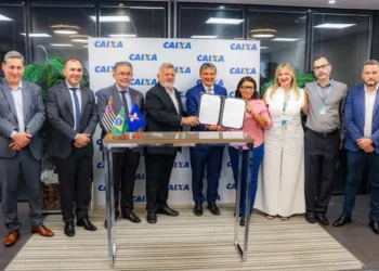 Está no CadÚnico? Veja como acessar o novo microcrédito de até R$ 21 mil 10 Grupo de pessoas formalmente vestidas segura contrato assinado em mesa com bandeiras, painel CAIXA ao fundo.