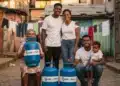 Inscrição no CadÚnico garante o recebimento do Gás do Povo? Entenda 4 Família sorridente com botijões do Gás do Povo em rua de comunidade.