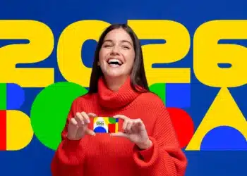 Mulher com suéter vermelho segura cartão do Bolsa Família sorrindo em frente a painel colorido com 2026.