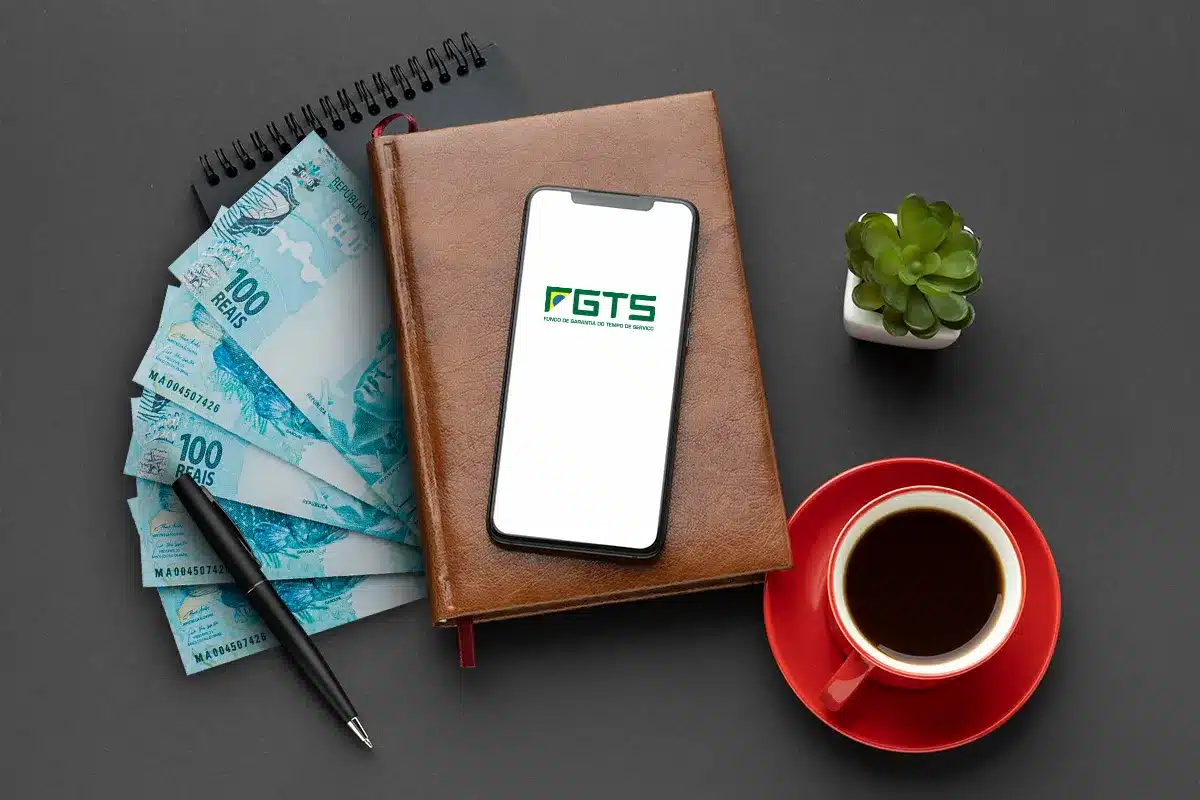Cédulas de R$ 100 e aplicativo FGTS em ambiente de trabalho Celular com app FGTS sobre caderno ao lado de notas de R$ 100, caneta, xícara de café e planta