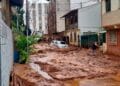 Rua coberta por lama após enchente com carro parcialmente submerso e homem caminhando em meio à destruição