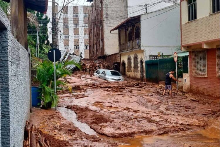 Rua coberta por lama após enchente com carro parcialmente submerso e homem caminhando em meio à destruição