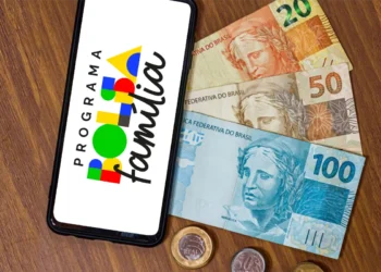 Celular com a logomarca do Programa Bolsa Família exibida na tela, ao lado de notas e moedas de reais sobre uma mesa, representando a consulta ao pagamento antecipado do benefício pelo aplicativo em abril de 2026