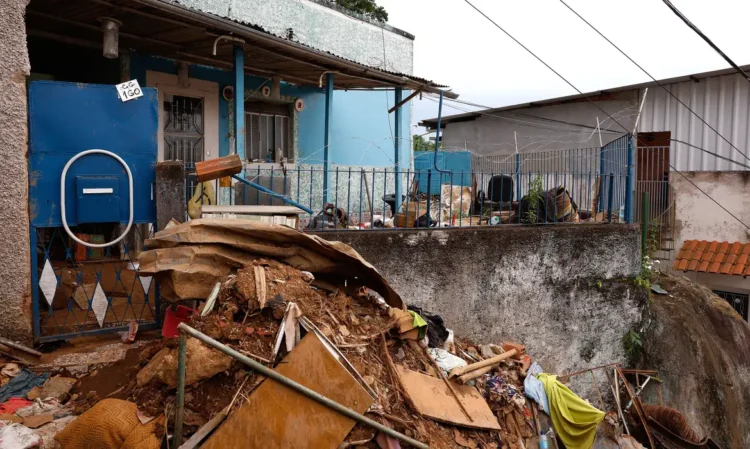 Auxílio Reconstrução MG 2026 Residência azul com escombros de lama e destroços acumulados após deslizamento de terra em área urbana brasileira.