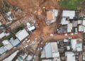 Vista aérea de deslizamento de terra atingindo casas em bairro residencial com escavadeiras e equipes de resgate no local.