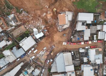 Vista aérea de deslizamento de terra atingindo casas em bairro residencial com escavadeiras e equipes de resgate no local.