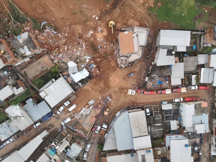 Vista aérea de deslizamento de terra atingindo casas em bairro residencial com escavadeiras e equipes de resgate no local.