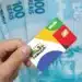 Bolsa Família: estar no CadÚnico não garante o benefício — entenda a regra 11 Mão segurando cartão colorido do Bolsa Família com notas de 100 reais ao fundo