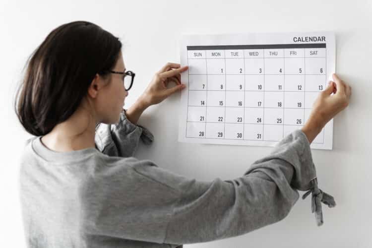 Jovem mulher de cabelos escuros com óculos pendura calendário grande com dias do mês na parede branca