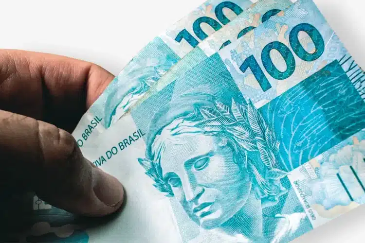Nova regra do IR pode colocar dinheiro na sua conta automaticamente 2 Mão segurando três notas de 100 reais brasileiros com efígie da República em destaque.
