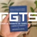 FGTS poderá ser utilizado em renegociações pelo Desenrola Brasil 2.0 12 Mão segura carteira de trabalho azul com logotipo FGTS em destaque e plantas verdes ao fundo.