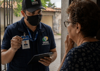 Homem com uniforme e crachá do INSS conversando com idosa em porta de casa