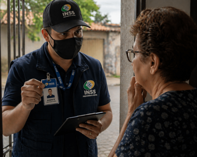 Homem com uniforme e crachá do INSS conversando com idosa em porta de casa