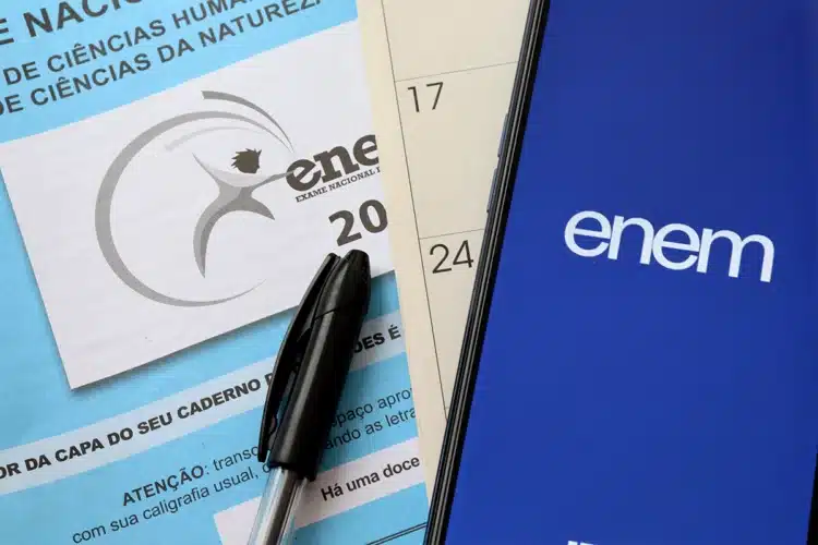 Enem 2026: participantes do Pé-de-Meia têm direito à isenção da taxa 2 Caderno de prova do Enem de Ciências Humanas e Natureza com caneta preta e smartphone mostrando logo do Enem azul.