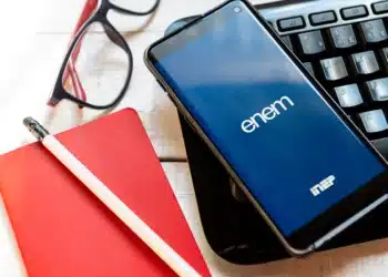 Celular com app Enem e logo INEP sobre teclado preto, caderno vermelho, óculos e lápis em mesa de madeira.