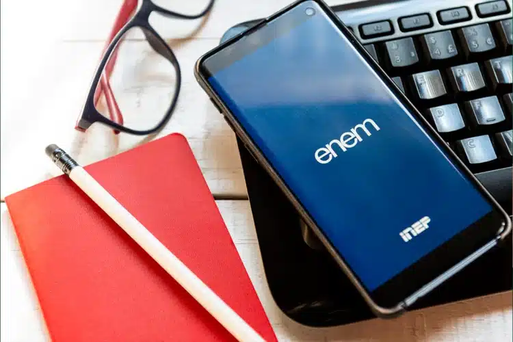 Enem 2026: participantes do Pé-de-Meia têm direito à isenção da taxa 1 Celular com app Enem e logo INEP sobre teclado preto, caderno vermelho, óculos e lápis em mesa de madeira.
