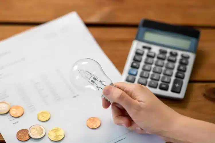 Conta de luz pode cair ou zerar? Governo envia mensagens e revela quem tem direito 2 Mão segura lâmpada incandescente sobre documento de conta com moedas de euro e calculadora ao fundo.