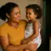 Mães de Pernambuco paga R$ 300 no dia 8 de abril; veja quem recebe 11 Mulher de camiseta amarela sorri segurando menina de vestido floral colorido em uma casa modesta