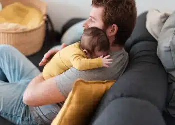 Homem sentado no sofá abraça bebê de roupa amarela sobre o ombro, almofadas ao redor