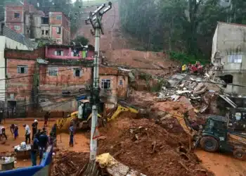 Receber o Auxílio Reconstrução de R$ 7,3 mil não afeta Bolsa Família ou BPC 7 Retroescavadeiras amarelas removem lama de deslizamento em comunidade com casas de tijolos vermelhos enquanto moradores observam.