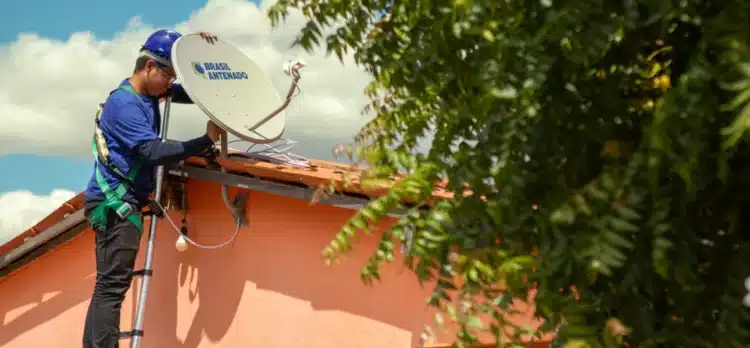 Programa Brasil Antenado: distribuição de antenas digitais gratuitas avança no país e amplia acesso à TV aberta 1 Técnico com capacete azul instala antena parabólica digital do programa Brasil Antenado no telhado de casa cor-de-rosa.