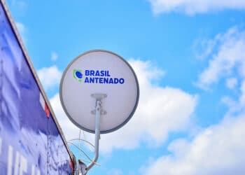 Antena parabólica digital branca com logotipo "Brasil Antenado" instalada em estrutura azul contra céu azul com nuvens.