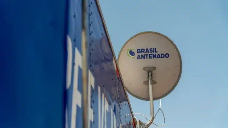 Programa Brasil Antenado: distribuição de antenas digitais gratuitas avança no país e amplia acesso à TV aberta 2 Antena parabólica digital branca com logo Brasil Antenado fixada em parede azul, céu claro ao fundo.