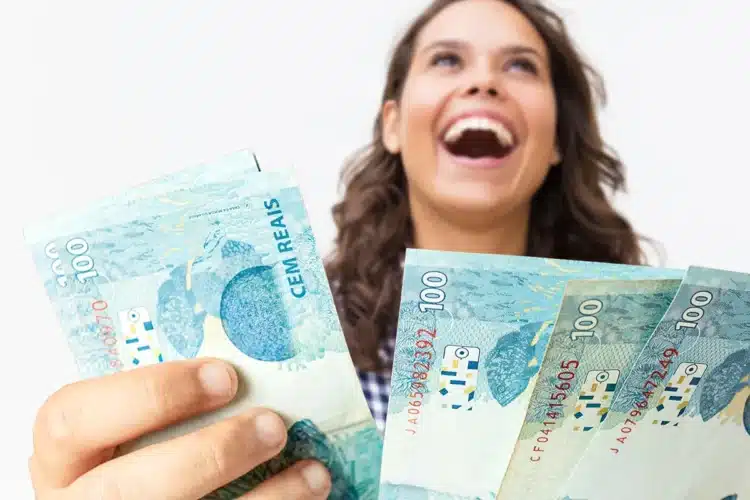 Salário mínimo de 2027 já foi definido? Veja tudo o que já se sabe 1 Mulher sorridente de cabelos castanhos ondulados segura quatro notas de R$ 100 em leque sobre fundo branco.