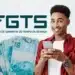 Aniversariantes de maio já têm data para sacar FGTS; confira calendário 12 Jovem de camisa vermelha segura smartphone sorrindo, com notas de R$100 e logotipo do FGTS ao fundo.