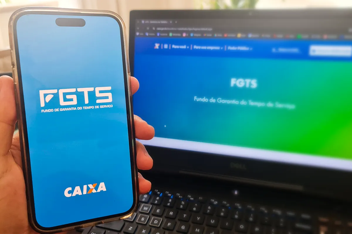 Saque-aniversário do FGTS é liberado nesta segunda-feira (4); descubra se você tem direito 2 Mão segurando celular com aplicativo do FGTS aberto ao lado de notebook exibindo o site oficial do Fundo de Garantia, ilustrando o saque-aniversário liberado em maio de 2026