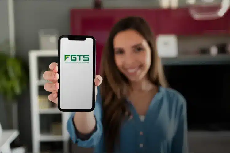 Saque-aniversário vs. saque-rescisão do FGTS: qual é a melhor opção para você? 2 Mulher sorridente de camisa azul mostra smartphone com logotipo do FGTS na tela, ambiente doméstico ao fundo.