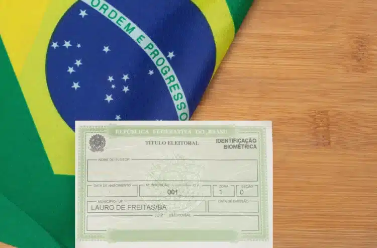 Situação eleitoral: como saber se seu título está apto para votar em 2026 2 Título de eleitor do município de Lauro de Freitas/BA posicionado sobre a bandeira do Brasil em superfície de madeira.