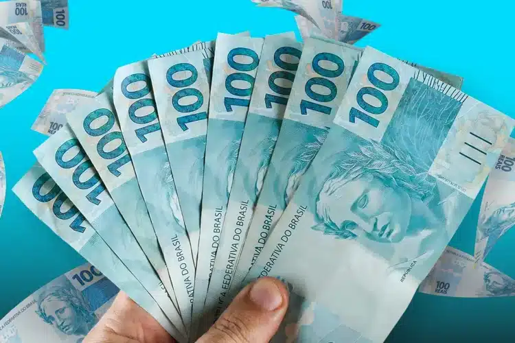 Mão segurando leque de notas de R$ 100 com fundo azul claro e cédulas espalhadas.