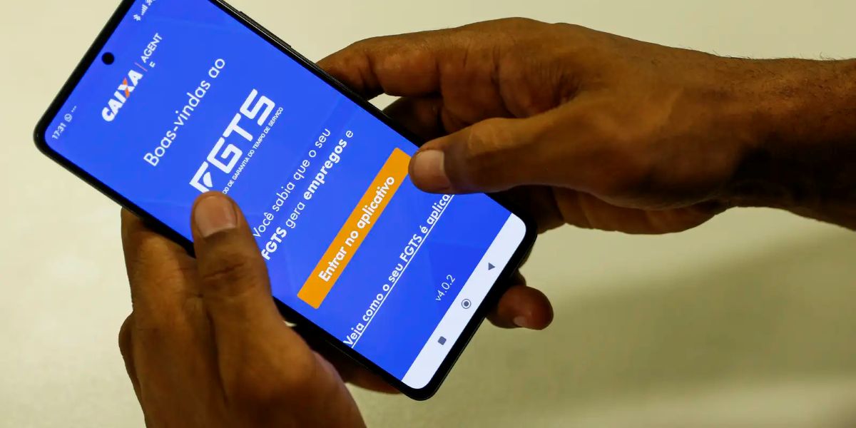 Trabalhadores demitidos de 2020 a 2025 recebem saque-aniversário: valores acima de R$ 3 mil só em agências 2 Mãos seguram um celular aberto no aplicativo FGTS da Caixa, na tela de boas-vindas