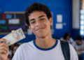 Enem: a regra que dá R$ 200 e isenção total para o estudante do Pé-de-Meia 12 Estudante de uniforme escolar sorri segurando uma nota de R$ 200 dentro de uma sala de aula